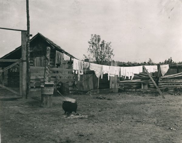 Clothing hanging on a line to dry in a farm yard. A pot of steaming water is in the foreground. A small open-sided farm building is behind a fence with a gate in the background.