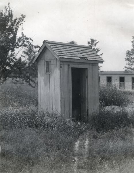 View of the front and side of an outhouse with an open door. A section of a long building with a row of windows is in the background.