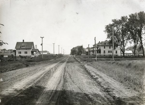 View down center of rural dirt road with houses along both sides in Black Hawk County. A man and horse are in a field on the left, near a sign that says: "Orange Cemetery."