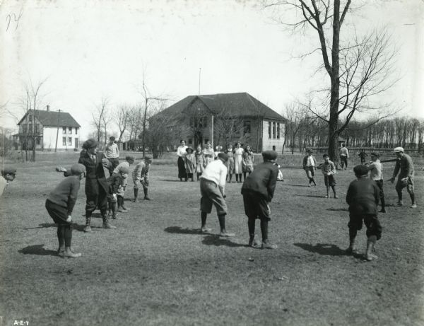 Children standing in a schoolyard playing baseball. Two buildings are in the far background.