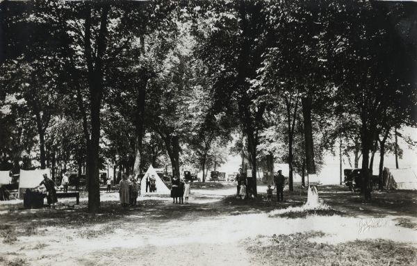 View down dirt road of people standing beneath the trees at Ottawa Park Tourist Camp. Automobiles are parked in the background, and tents are set up throughout the clearing.