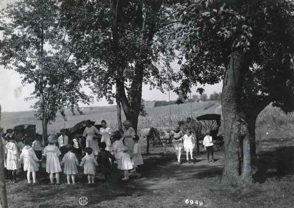 A group of children are standing in a circle under trees while playing an outdoor game led by Mrs. Markley. Two children are sitting on a tree swing to the right, and horses and a horse-drawn carriage are in the background.