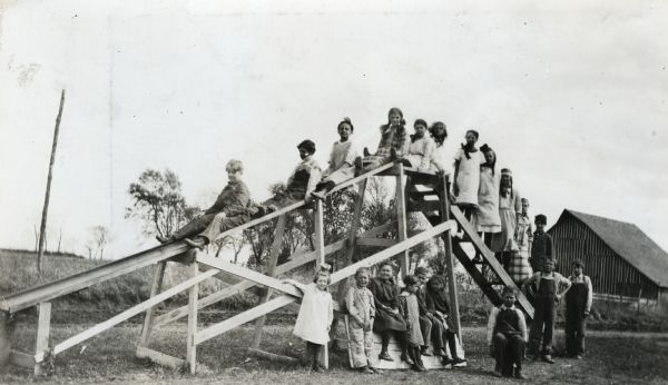 A group of children posing on and beneath a wooden playground slide. There is a barn in the background.