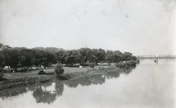 Elevated view over the Illinois River towards Allen Park, a free tourist camp. Tents are set up along the bank of the river, and a bridge spans the water in the background.