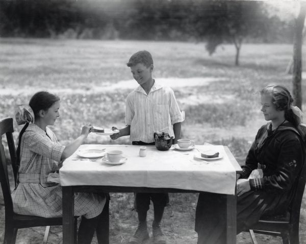 Two girls are sitting at a table outdoors while a boy is offering them sandwiches from a plate during an etiquette lesson.