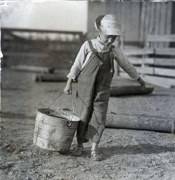 A boy wearing overalls and a straw hat is carrying a metal pail across a barnyard.