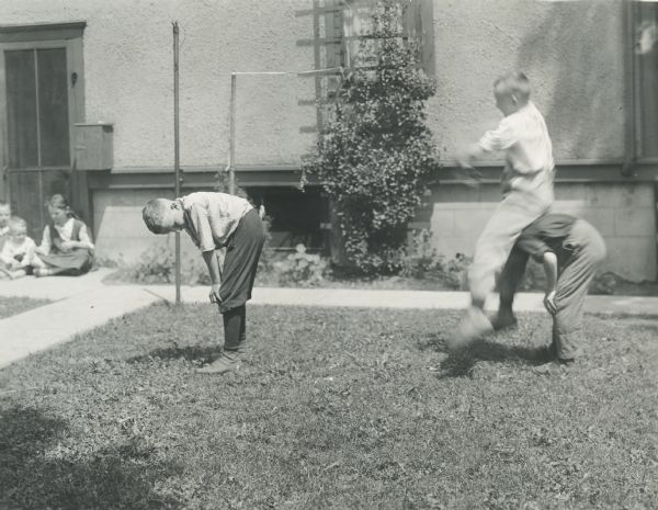 Boys from Sunnyside School playing leapfrog on the lawn outside a building. Other children are sitting near a screen door in the background.