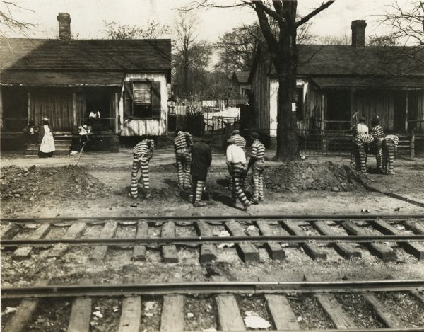 A group of convict laborers are wearing striped jumpsuits as they work to construct a road. Railroad tracks run through the foreground and people are standing near homes in the background.