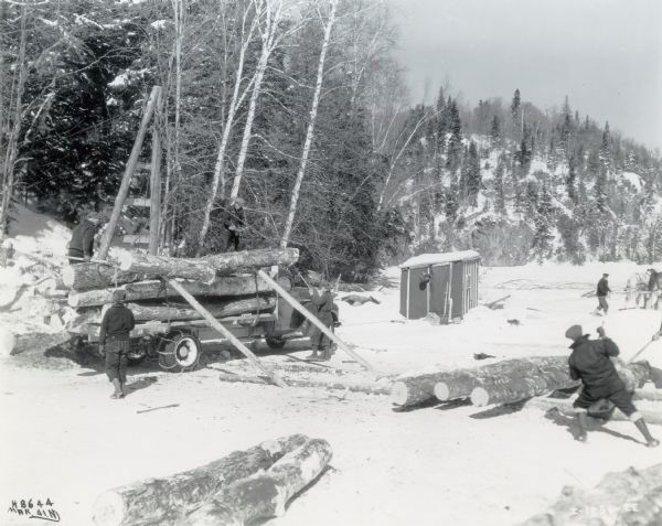 Men use two pieces of lumber to unload or load cut logs from the back of a truck outfitted with snow chains near Blairhampton in Ontario, Canada. On the right two men are working with logs on the ground. A small building is in the background, along with a man leading a team of horses.