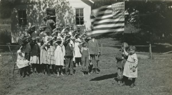 A boy and a girl are holding an American flag while standing in front of a building, possibly a schoolhouse. A group of children is standing in a triangle formation nearby while saluting the flag.