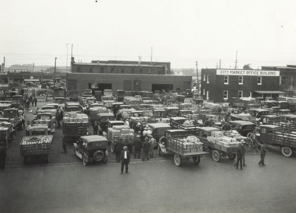 Elevated view of men standing around vendor trucks parked at the city fruit market in front of commercial buildings. A building in the background has a sign reading: "City Market Office Building."