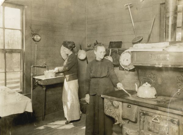 Miss Foy Martin filling a bucket with water from a sink faucet, while her mother is lighting a burner on the stove inside the kitchen of R. Martin.