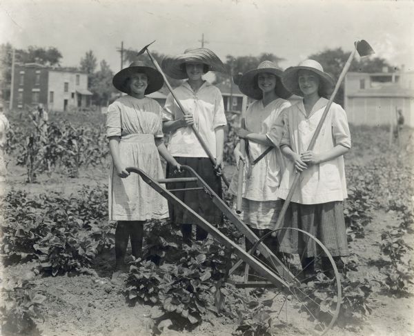 Group portrait of women wearing wide-brimmed hats standing outdoors while holding hoes and a garden plow in what appears to be a city garden. Several buildings are in the background.