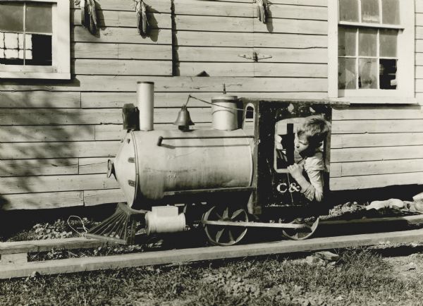 View across years towards a boy looking out the window of a cab of a toy locomotive as he is moving along a set of wooden tracks. Behind him is the wall of a building from which bundles of dry corn are hanging.