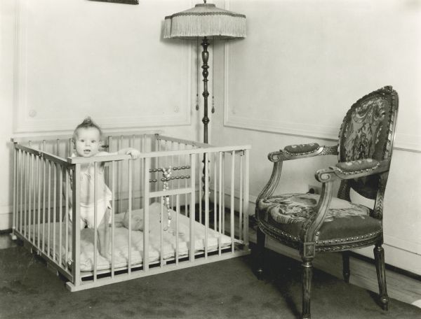 An infant wearing a diaper standing and holding onto the rails of a playpen in the corner of a room. A lamp and an upholstered chair are nearby. Toy beads and a stuffed animal are inside the playpen.