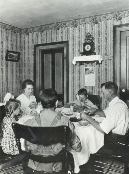 A family with three children sitting around a dining table while eating a meal. The walls of the dining room are covered with wallpaper and decorated with a framed picture, clock, and calendar.