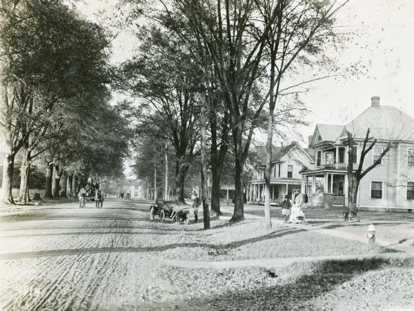 View down dirt-covered residential street among trees. A horse-drawn carriage is coming down the road, and a woman and child are standing on the sidewalk on the right. Two boys are standing near an automobile parked along the side of the road.