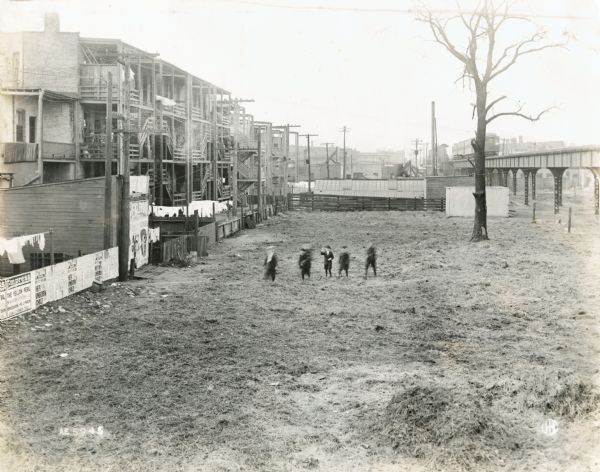 Elevated view of five children walking in a field behind what appears to be tenement housing. Wooden fire escapes are attached to the rear of the buildings, and an elevated train passes by along tracks to the right. Signs on the buildings and fences advertise what appear to be theater shows entitled: "The Yellow Peril" and "Her Unborn Child."