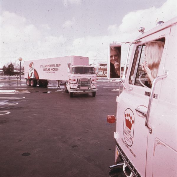 A "Cameo Girl," saleswoman for the Cameo Retail Division of Bestline Incorporated, sitting in the cab and applying lipstick while looking in the driver-side mirror of a pink-colored International Transtar C-O-F4070A diesel truck outfitted with a 40-foot trailer. Another truck is in the background, with text on the exterior of the trailer reading: "It's a Wonderful New Bestline World; Retail Cameo Division."