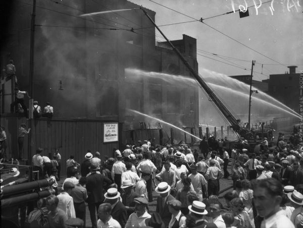 Elevated view of a crowd gathered outside International Harvester's Deering Works at Fullerton and River, as a crew of firemen works to extinguish a fire inside the factory building. The sign on the fence reads, "For Sale or Lease on Switch Will Divide. J.H. Van Vlissingen & Co. 120 S. LaSalle St. Randolph 4042."