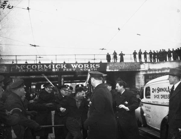 Police officer and marchers from a Congress of Industrial Organizations (also known as the Committee of Industrial Organization) union clash outside International Harvester's McCormick Works factory during a worker strike. Men watch the fight from a bridge or walkway in the background. The man in the center of the photograph with a clenched fist is Ruben Silverman.