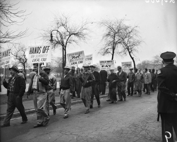 Factory workers hold signs as they picket while participating in a strike. A uniformed police officer looks on from the right. The workers are picketing near International Harvester's Tractor Works.