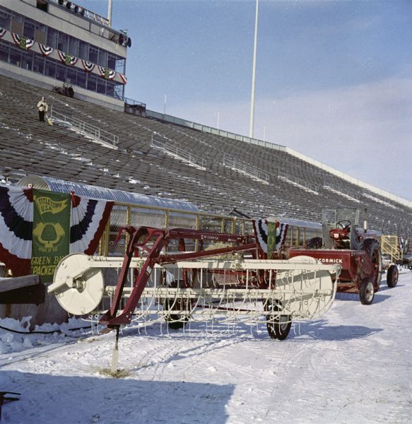 Color photograph of a Farmall tractor and hay rake on the sideline at City Stadium (later known as Lambeau Field), home of the Green Bay Packers. The equipment was used to remove bales of hay that were protecting the frozen field before a game against the New York Giants. A man sweeping snow from the seats is in the background.