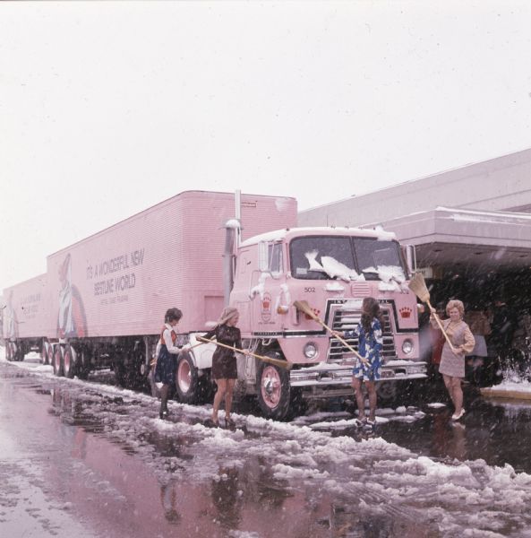 Four "Cameo Girls," saleswomen for the Cameo Retail Division of Bestline Incorporated, using brooms to sweep snow from a pink-colored International Transtar C-O-F4070A diesel truck outfitted with a 40-foot trailer. The text on the trailer reads: "It's a Wonderful New Bestline World; Retail Cameo Division."
