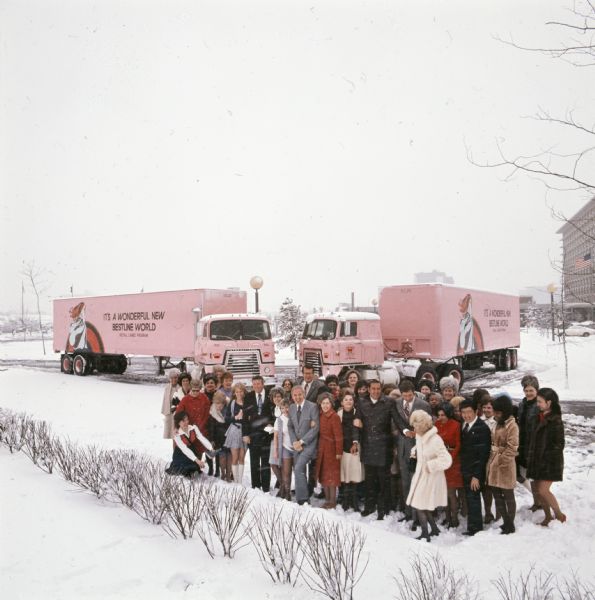 Elevated view of a group of salesmen and "Cameo Girl" saleswomen from Bestline Incorporated. They are standing in a snow-covered parking lot in front of two pink-colored International Transtar C-O-F4070A diesel trucks outfitted with 40-foot trailers. The text on the trailers reads, "It's a Wonderful New Bestline World; Retail Cameo Division."