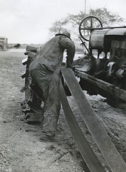 A man putting a tractor belt on a pulley while the machine is in operation, demonstrating a farm hazard. Taken at International Harvester's Hinsdale experimental farm.