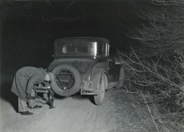 A man is standing alongside a dirt road while changing a rear tire on an automobile, blocking the taillight and demonstrating a farm hazard.