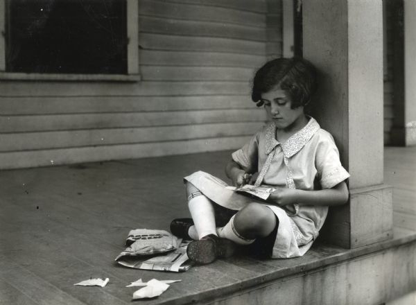 A girl is sitting and leaning on a porch column outdoors while using a scissors to clip something from a newspaper. Taken at International Harvester's Hinsdale experimental farm to demonstrate the hazards of allowing children to use sharp scissors.