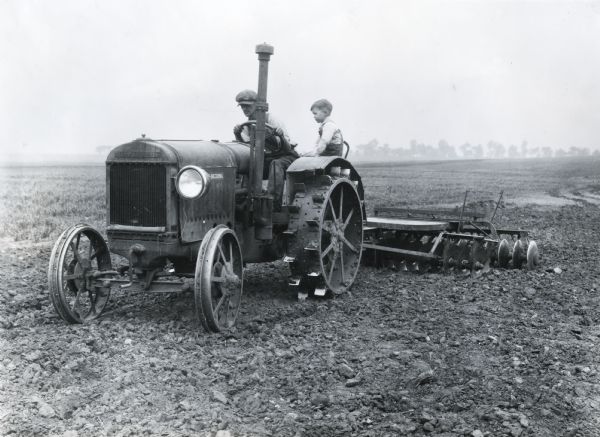 A child is riding on the fender of a tractor as a man is driving it through a field on International Harvester's Hinsdale experimental farm, demonstrating a farm hazard. The tractor is equipped with a large spotlight.