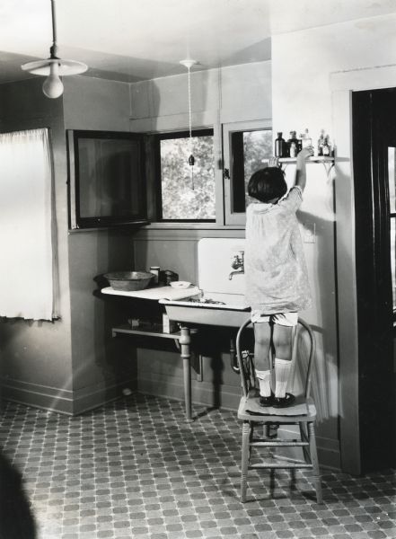 A girl is standing on a chair in the kitchen at International Harvester's Hinsdale experimental farm in order to reach a shelf where bottles of poison are kept. The photograph was staged to demonstrate a farm hazard.