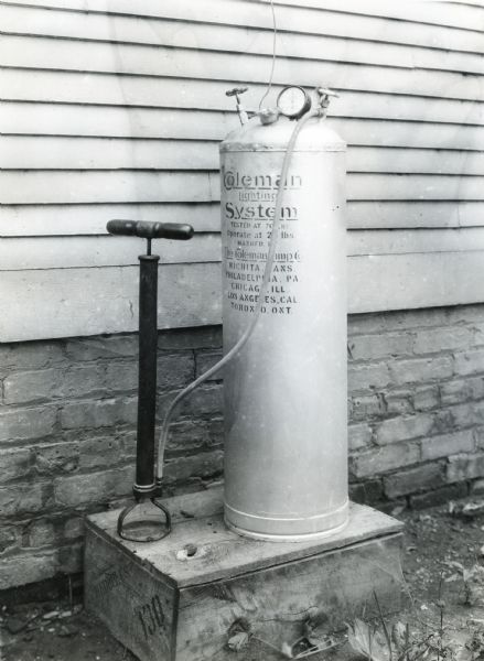 View of a pressure tank for a Coleman Air Gas stove placed on a wooden crate outside of a building on International Harvester's Hinsdale experimental farm, demonstrating a farm hazard.