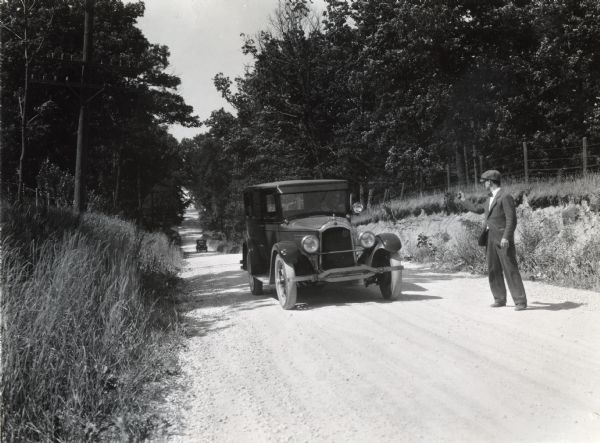 A hitchhiker thumbing a ride from a passing car along a gravel road near Hinsdale. The photograph was staged to demonstrate a farm hazard.