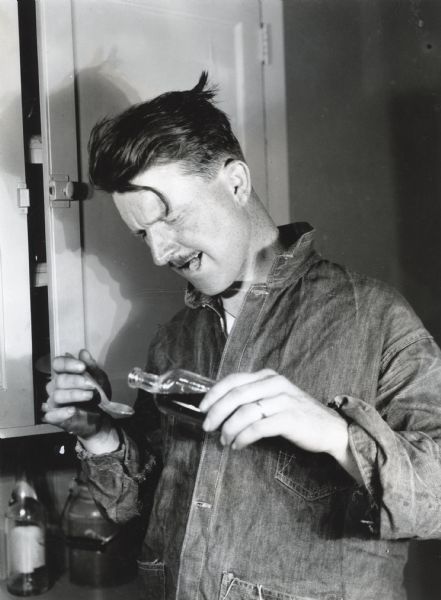 A man holding a cork between his teeth is pouring a dose of medicine into a spoon from an unmarked bottle. The photograph was staged at International Harvester's Hinsdale experimental farm to demonstrate a farm hazard.