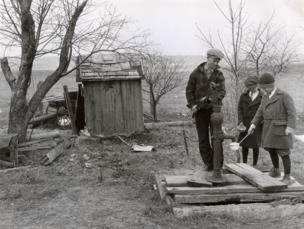 A boy wearing a winter coat and hat is pumping water from a well into ladles two other boys are holding. A shed, wheelbarrow, and wood scraps are in the background. The photograph was taken to demonstrate the farm hazard of a leaky well top.