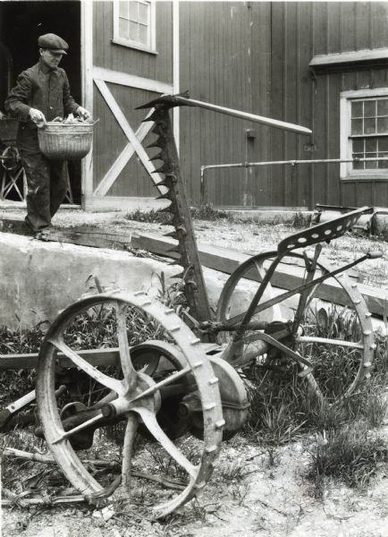 A mowing machine with its cutter raised is resting near the entrance to a barn as a man is walking nearby while carrying a metal bucket. The photograph was staged on International Harvester's Hinsdale experimental farm to demonstrate a farm hazard.