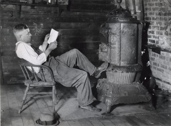 A man is sitting in a wooden chair while reading and resting his foot against a woodstove. The photograph was staged at International Harvester's Hinsdale experimental farm to demonstrate the danger of sitting near a stove during a thunderstorm.