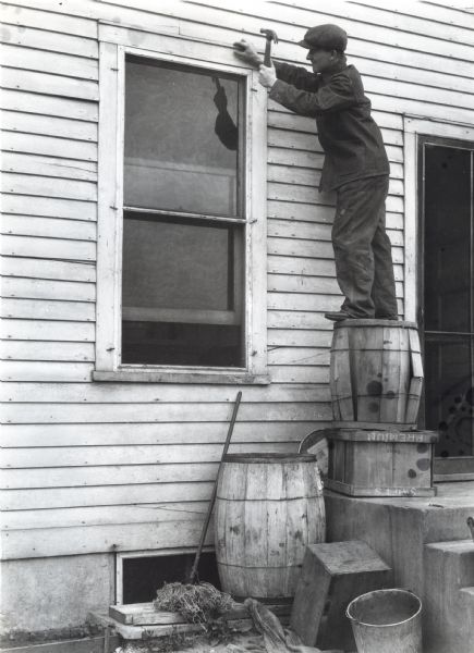 A man is standing on a wooden barrel which is resting on a wooden box on concrete entrance steps near a door. He is using a hammer to fix a window frame. The photograph was staged at International Harvester's Hinsdale experimental farm to demonstrate a farm hazard.
