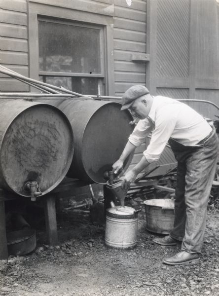 A man is using a funnel to siphon gasoline from a tank into a pail while smoking a pipe. The photograph was staged on the K. Legge Memorial Farm to demonstrate a farm hazard.