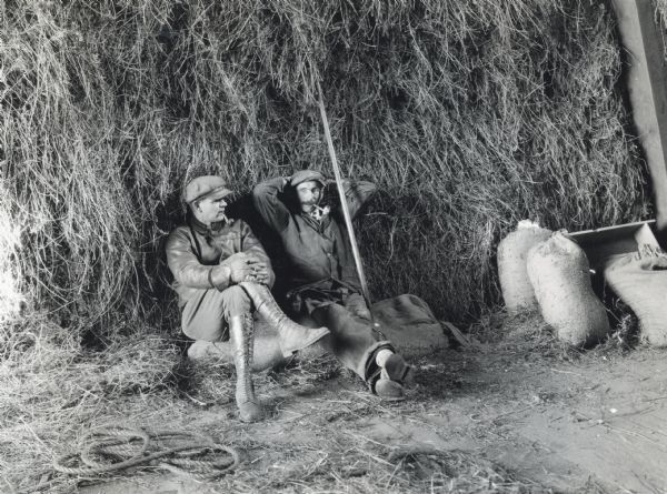 Two men are resting against a haystack while smoking pipes. The photograph was staged at International Harvester's Hinsdale experimental farm to demonstrate a farm hazard.