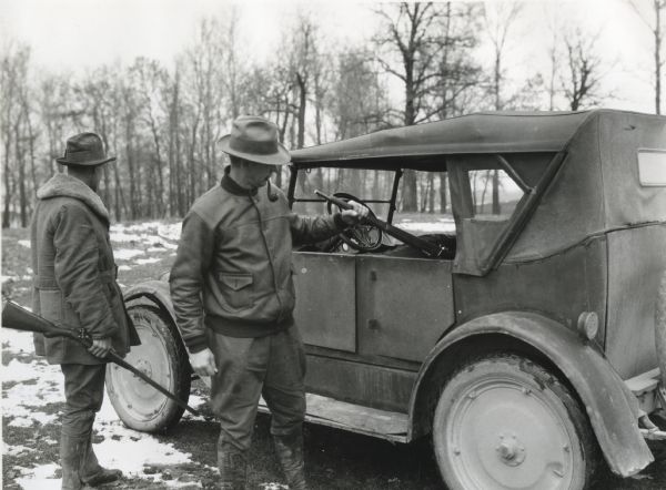 A man smoking a pipe is pulling a gun by the muzzle out from the back seat of an automobile, while another man holding a gun is standing beside him. The scene was staged on International Harvester's Hinsdale experimental farm to demonstrate a farm hazard.