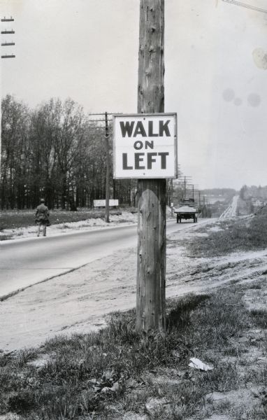 A sign reading: "Walk on Left" is posted on a power line pole. A man is walking on the left side of a rural road in the background as a truck drives by.