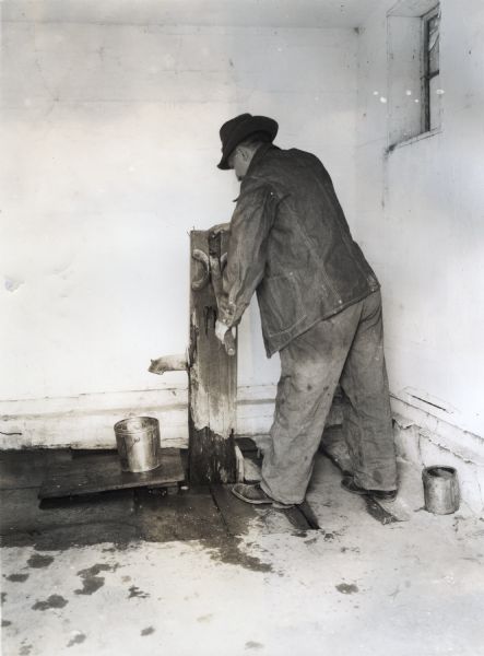A man using a wooden pump to fill a metal pail with water. The photograph was staged to demonstrate a hazard found on farms.