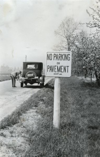 A sign reading: "No Parking on Pavement - State Law" is posted alongside a road, and in the background a man is standing in the line of traffic to inspect the tire of his automobile in the background.