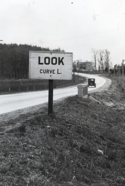 A sign reading: "Look Curve L." is posted along the side of a paved road. An automobile is on the road in the background.