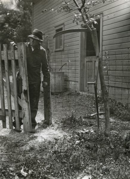 A man is working to fix a wooden fence while standing near a scythe which is hanging from the branch of a nearby tree, demonstrating a farm hazard. A farm building is in the background.