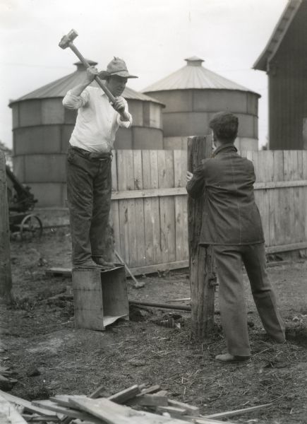 A man is standing on a crate while using a sledge hammer to strike a piece of wood being held in place by another man who is standing in front of him. Two silos and a barn are in the background.